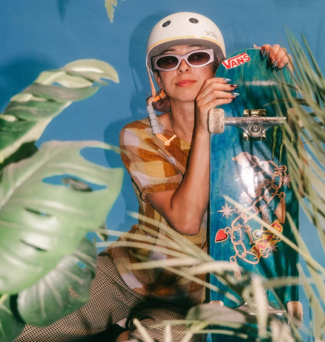Skateboarder with white helmet and white sunglasses holds a blue longboard with stickers against a bright blue backdrop.
