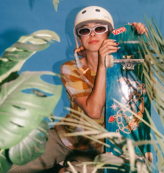 Skateboarder with white helmet and white sunglasses holds a blue longboard with stickers against a bright blue backdrop.